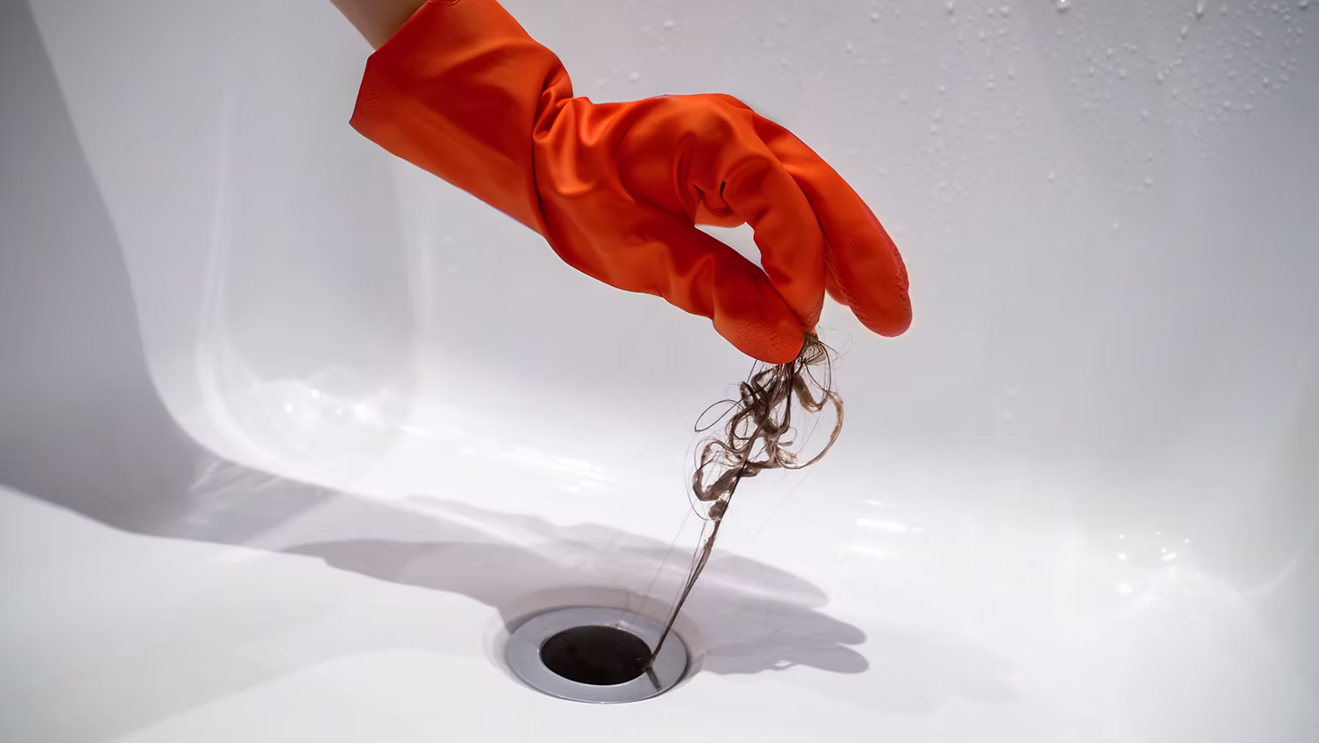 Hand wearing an orange glove holding hair and debris extracted from a sink drain, illustrating the need for professional drain cleaning services to prevent plumbing issues.